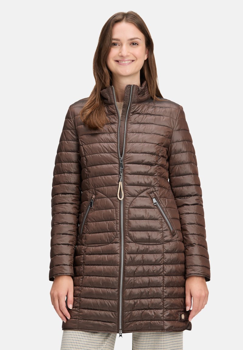 Smilende kvinde iført en brun quilted lang pufferjakke med lynlåslommer over lyse ternede bukser, stående mod en ensfarvet baggrund.