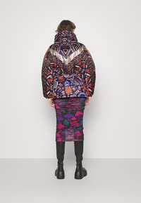 Personne portant une doudoune à motifs multicolores, une jupe noire avec des imprimés de champignons colorés, et des bottes à plateforme noires, vue de dos.