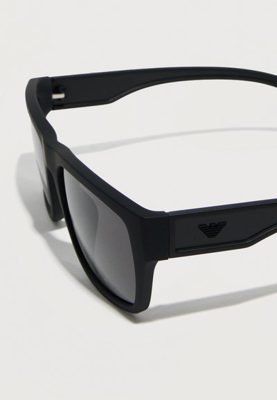 Μαύρα ορθογώνια γυαλιά ηλίου με παχιά πλαίσια και σκοτεινούς φακούς, με μικρό ανάγλυφο λογότυπο στον κλαδάκι του βραχίονα.