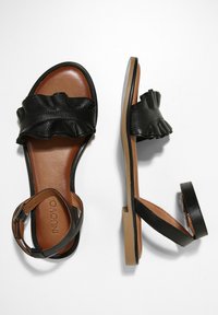 Czarne skórzane sandały z falbaniastym paskiem, brązową wkładką, płaską podeszwą i regulowanym paskiem na kostce. Prosty i swobodny styl.