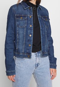 Tmavě modrá džínová bunda s dvěma náprsními kapsami, zapínáním na knoflíky a krátkým střihem. Nositelná přes černé tričko se zipem.