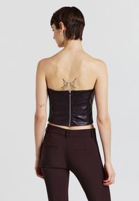 Corsetto senza spalline in pelle nera con cerniera posteriore e design aderente. Modello indossa pantaloni sartoriali marrone scuro.