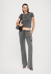 Persoon in een grijze cropped t-shirt en bijpassende wijde broek met een zwarte tas over de schouder en open teen schoenen, staande tegen een witte achtergrond.