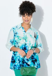 Blusa leggera con stampa floreale blu e verde, con bottoni e colletto, maniche a 3/4. Abbinata a una borsa metallica verde e accessori.