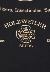 Černá tkanina s krémovým textem a grafikou. Obsahuje "HOLZWEILER" a "SEEDS" s květinovým logem označeným "QUALITY." Jednoduchý design.