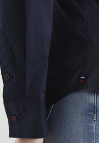 Granatowa koszula z długim rękawem z mankietami na guziki, gładka tekstura, zaokrąglony dół oraz subtelne czerwone i białe akcenty w pobliżu linii dolnej.