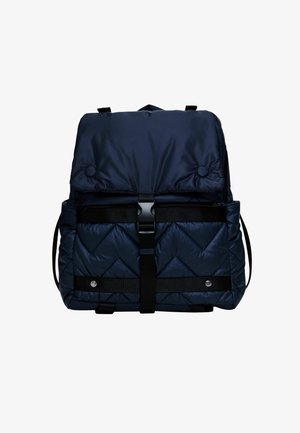 Mochila azul marino acolchada con cierre de solapa, correa con hebilla negra, bolsillos laterales y detalles de botones sobre un fondo blanco.