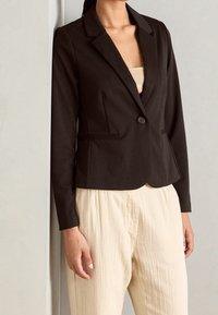 Černý přiléhavý blazer s výstřihy ve tvaru V, zapínáním na jeden knoflík a mírnou texturou. Spárován s lehkými, volně padnoucími béžovými kalhotami.