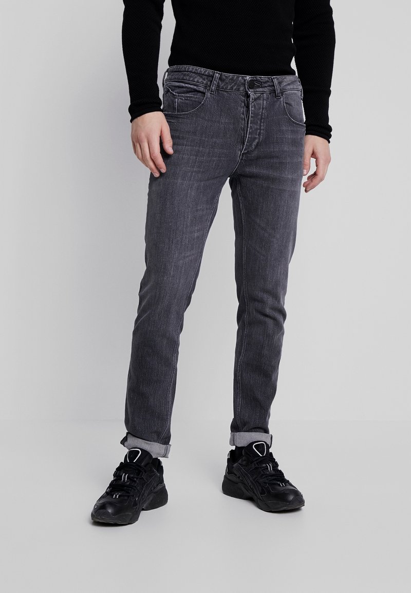 Vaqueros de mezclilla gris con un ajuste slim y textura desgastada, con trabillas para cinturón, bolsillos delanteros y dobladillos enrollados, combinados con zapatos deportivos negros.