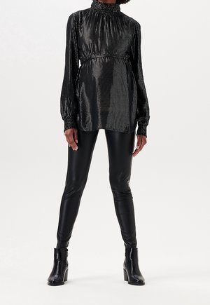 Osoba ubrana w błyszczącą czarną bluzkę z wysokim kołnierzem i długimi rękawami, czarne skórzane legginsy i czarne botki na białym tle.