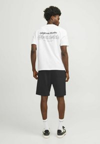 Vitt bomullströja med svart "Originals Studio Worldwide" grafisk design på ryggen, kombinerad med svarta shorts och sneakers, stående mot en enkel bakgrund.