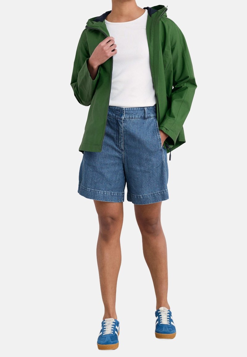 Green waterproof jacket, white ribbed tank top, blue denim shorts, blue sneakers with white stripes and tan soles. Casual outdoor outfit.