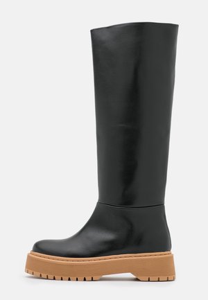 Botte haute en cuir noir avec semelle épaisse texturée couleur tan et bout arrondi, profil latéral sur fond gris clair.