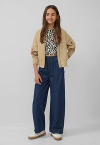 Beige Bomberjacke über einem schwarz-weißen Leopardenmuster-Crop-Top, kombiniert mit lockeren blauen Denim-Hosen und weißen Sneakers mit braunen Sohlen.