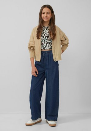 Beige bomberjack over een zwart-wit panterprint crop top, gecombineerd met losse blauwe denimbroek en witte sneakers met tan-zolen.