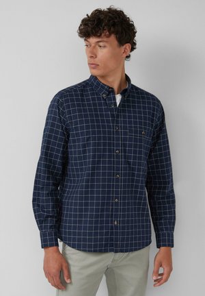 Chemise bleu marine à boutons avec un motif à carreaux en bleu clair et beige. Elle possède des manches longues, un col pointu et une poche poitrine.