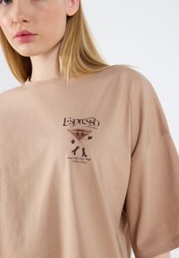 Camiseta de algodón beige con mangas cortas. Presenta un gráfico marrón de un personaje de copa de martini y texto que dice "Cóctel de Espresso."
