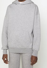 Personne portant un sweat à capuche gris clair et un pantalon de survêtement assorti, les mains détendues le long du corps sur un fond uni.
