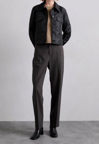 Zwarte cropped jack met twee borstzakken, beige trui en grijze op maat gemaakte broek. Zwarte enkellaarsjes met hak maken de outfit compleet.