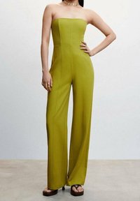 Žena v bezramínkovém širokém kombinézovém kostýmu v limetkové zelené barvě, s platformovými sandály, zlatým náhrdelníkem a s rukou opřenou o bok.
