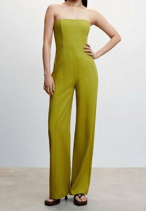 Femme portant une combinaison ample sans bretelles de couleur vert lime avec des sandales à plateforme, un collier en or et la main reposant sur la hanche.