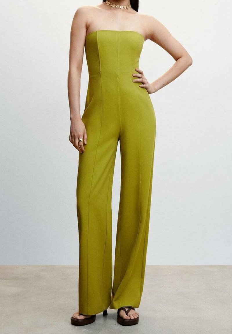 Žena v bezramínkovém širokém kombinézovém kostýmu v limetkové zelené barvě, s platformovými sandály, zlatým náhrdelníkem a s rukou opřenou o bok.