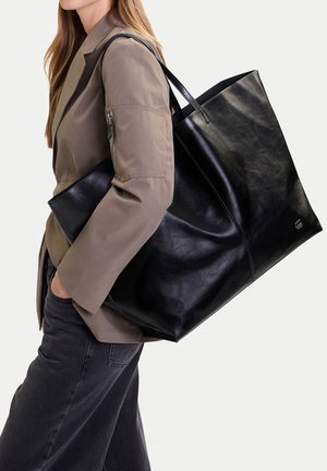 Mujer con blazer color topo y vaqueros negros, llevando un bolso tote de cuero negro de gran tamaño al hombro.