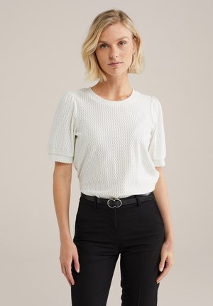 Haut blanc texturé à manches courtes avec un col rond, associé à un pantalon noir et une ceinture avec une boucle en argent circulaire.