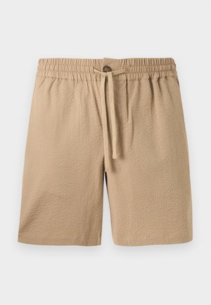 Homme portant une chemise beige à manches courtes avec des motifs marron, un short texturé couleur beige, des sandales noires et des lunettes de soleil foncées, debout les mains dans les poches.