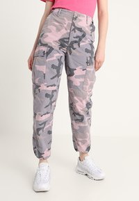 Kamouflage cargobyxor i grå och rosa nyanser, med elastiska mouser, flera fickor och en avslappnad passform, tillsammans med vita sneakers.
