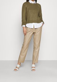 Oliwkowo-zielony dziergany sweter na białej koszuli z kołnierzykiem, w zestawieniu z beżowymi spodniami ze skóry ekologicznej i białymi sandałami na słupku.