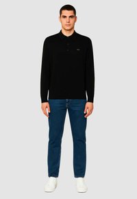 Polo nero a maniche lunghe con colletto, contrassegnato da un piccolo logo verde. Abbinato a jeans blu e sneakers bianche. Design semplice.