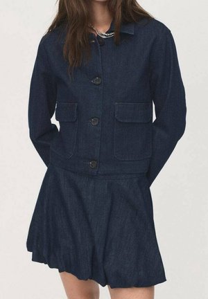 Femme portant une veste en denim bleu foncé avec de grandes poches avant et une jupe évasée assortie, debout les mains derrière le dos.