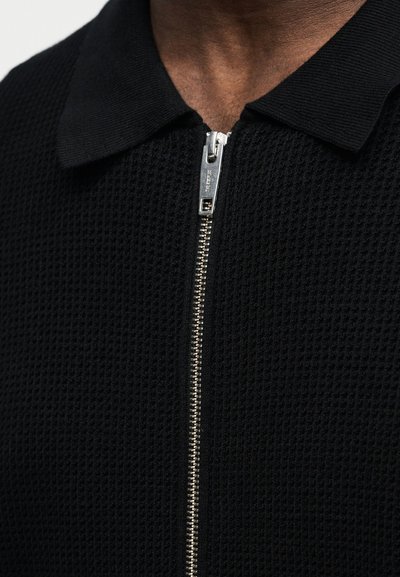 Κοντινό πλάνο ενός ατόμου που φοράει μαύρο πουκάμισο με υφή, με φερμουάρ ασημί και επίπεδη γιακά.