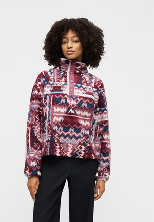 Mujer con cabello rizado que lleva un pulóver de forro polar con estampado multicolor y botones de presión, y pantalones negros, de pie frente a un fondo liso.
