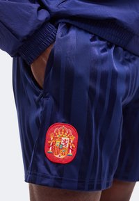 Main dans la poche, portant un short bleu marine avec des rayures verticales foncées et un écusson rouge des armoiries espagnoles sur la cuisse gauche.