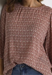 Haut couleur rouille avec un motif de feuilles. Le tissu est léger avec une texture agréable. Encolure ronde et manches amples et fluides.