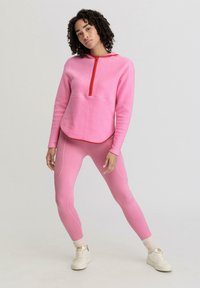 Maglione in pile rosa con zipper rosso e rifiniture, abbinato a leggings rosa con dettagli sulle cuciture. Scarpe da ginnastica bianche completano l'outfit.