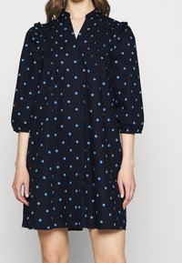 Marinblå klänning med blåa prickar, volangslyft vid axlarna, en V-ringning och samlade veck på framsidan. Knäppning synlig.