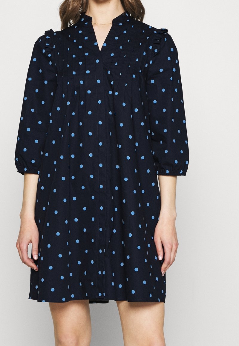 Marinblå klänning med blåa prickar, volangslyft vid axlarna, en V-ringning och samlade veck på framsidan. Knäppning synlig.