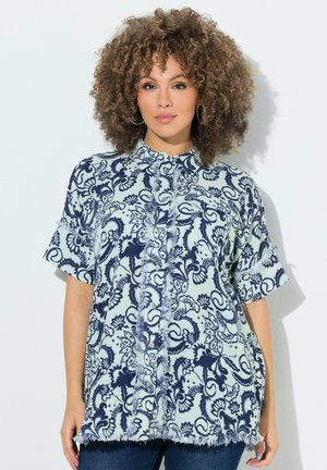 Kvinde med krøllet hår iført en blåt og hvid blomsterprintet kortærmet bluse med flæsekant og blå jeans foran en hvid baggrund.