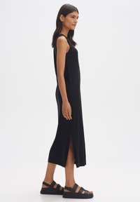 Zwarte mouwloze jurk met zijsplitten, gemaakt van een glad materiaal. Gepaard met zwarte sandalen met bandjes en dikke zolen.