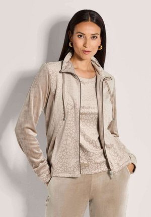 Frau trägt eine beige Samt-Jacken mit Reißverschluss, dazu passende Bluse und Hose, posiert mit den Händen in den Taschen vor einem schlichten Hintergrund.