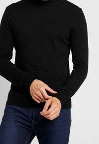 Svart stickad höghalssweater med långa ärmar och ribbade muddar. Tyget ser mjukt och texturerat ut, kombinerat med mörka denimjeans.