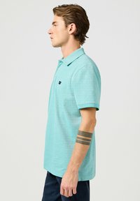 Polo shirt turchese chiaro con colletto, patta con due bottoni e un piccolo logo sul petto. Tessuto morbido con maniche corte e taglio dritto.