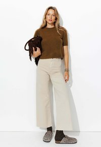 Pull marron court, pantalons larges beiges, sabots imprimé animal, et sac à franges marron. Cheveux longs et bracelets en argent comme accessoires.