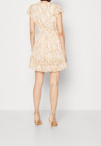 Femme portant une robe courte en imprimé serpent beige avec un ourlet volé et des manches flottantes, associée à des sandales à talons nude, se tenant de dos.