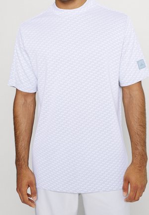 Homme portant une chemise blanche à manches courtes à motifs avec un petit patch du logo Adidas sur la manche gauche.