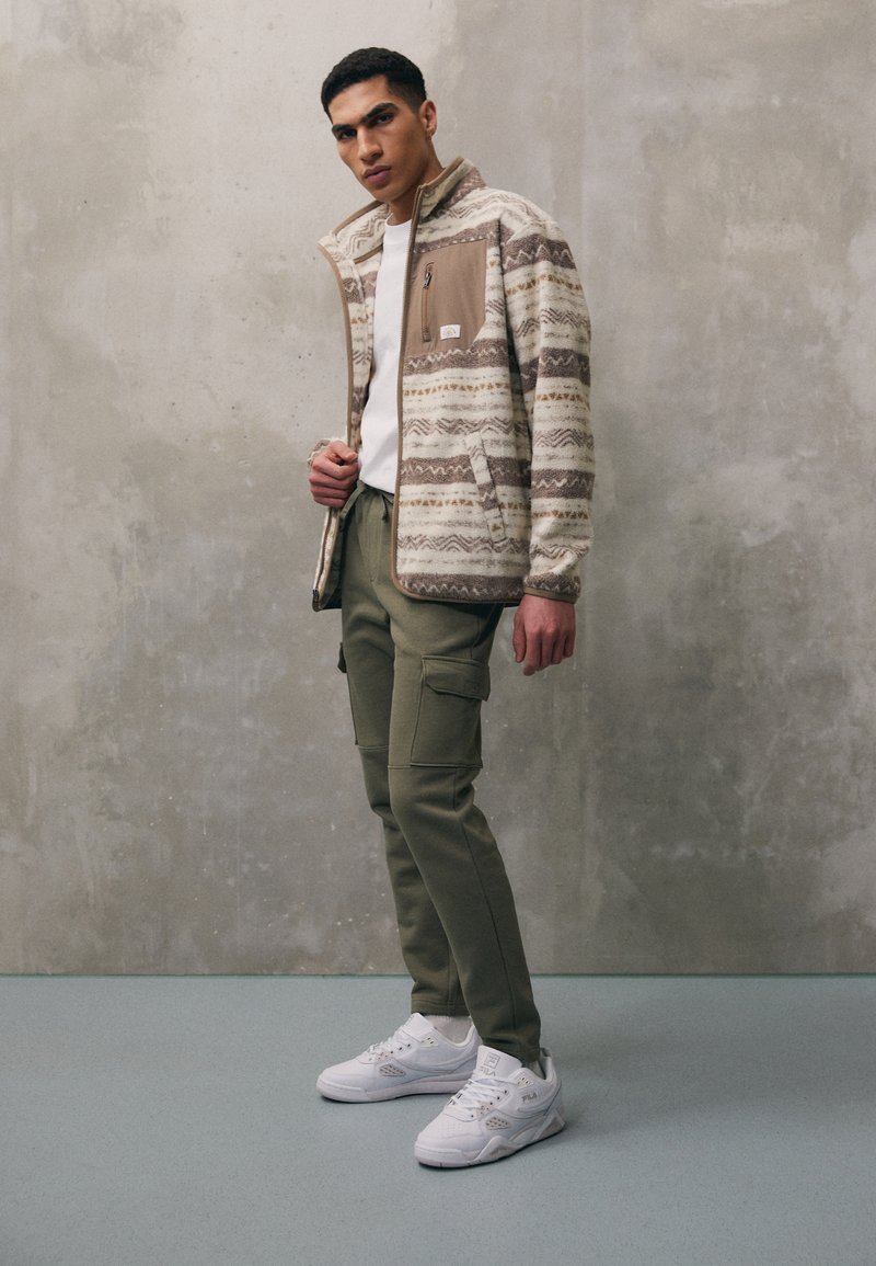 Fleecejacke mit geometrischem Muster in Beige und Braun, ausgestattet mit einer braunen Reißverschlusstasche; kombiniert mit olivgrünen Cargo-Hosen und weißen Sneakers.