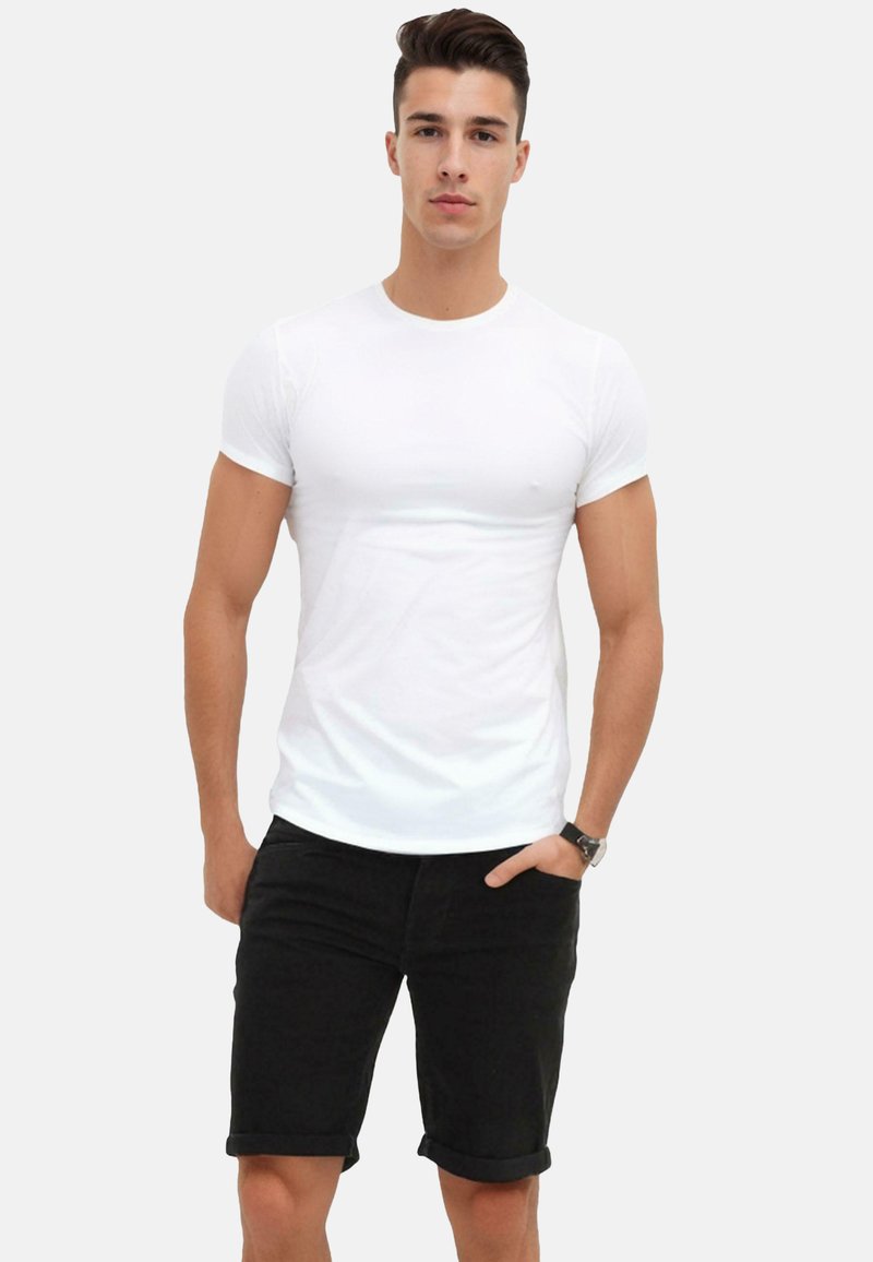 White, short-sleeve t-shirt with a classic crew neckline, paired with black rolled-up denim shorts. Model standing with hand in pocket.